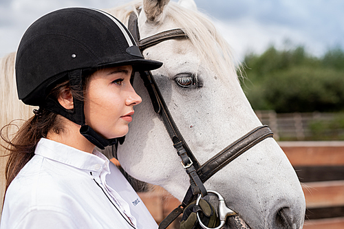 young calm female in white shirt and equestrian helmet standing by racehorse in rural