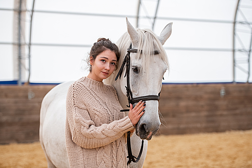 Pretty young brunette active woman in casual sweater standing close to white horse and touching her muzzle