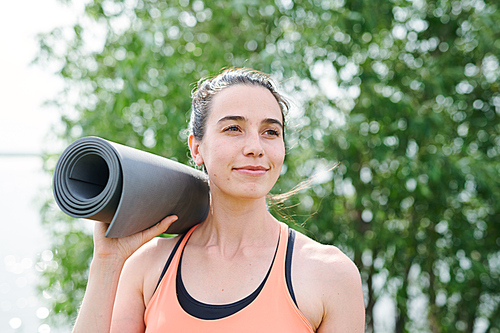 Content purposeful yoga trainer holding exercise mat on shoulder and looking into distance outdoors