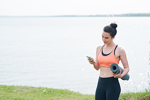 Smiling beautiful young woman standing against lake and holding exercise mat while checking yoga plan on phone