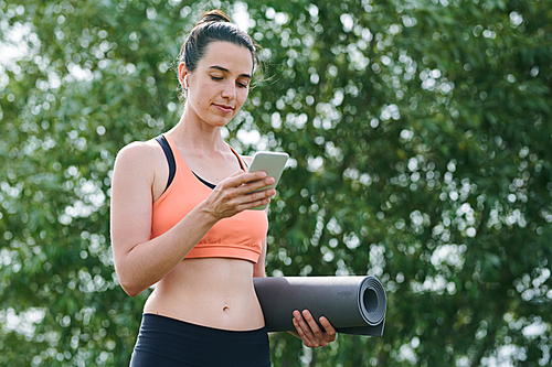 Content pretty woman in sports bra standing against tree in park and using phone while preparing for yoga practice outdoors