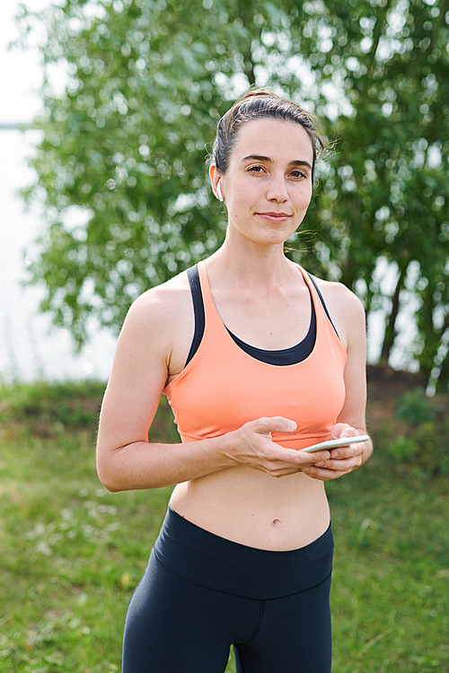 Portrait of content sporty young woman in sportswear standing in park and using modern gadget