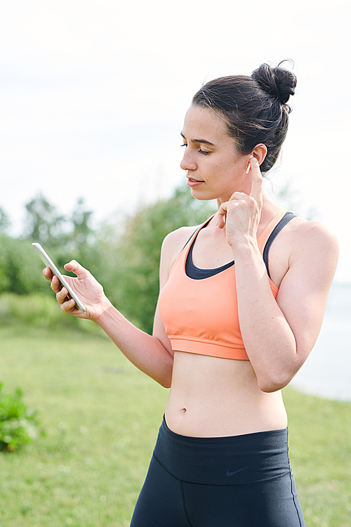 Serious young brunette woman adjusting wireless earphones while choosing music for yoga training outdoors