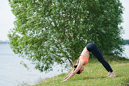 Flexible girl in sportswear performing downward facing dog on shore in summer