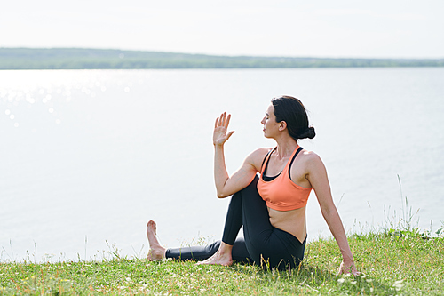 Calm young woman in sports clothing sitting on grass at lake and keeping eyes closed while twisting spine outdoors