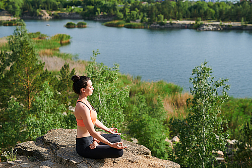 Young woman in sports clothing sitting in lotus position and doing breathing exercise outdoors
