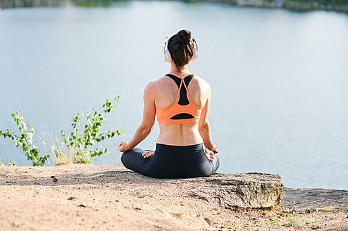 Rear view of young woman in wireless headphones sitting in lotus position and meditating with music outdoors
