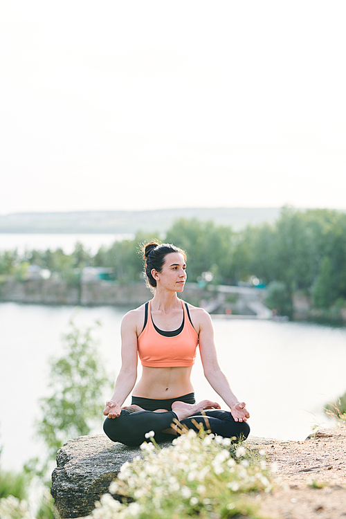 Content female yogis in sports bra sitting in lotus position and contemplating nature while enjoying meditation outdoors
