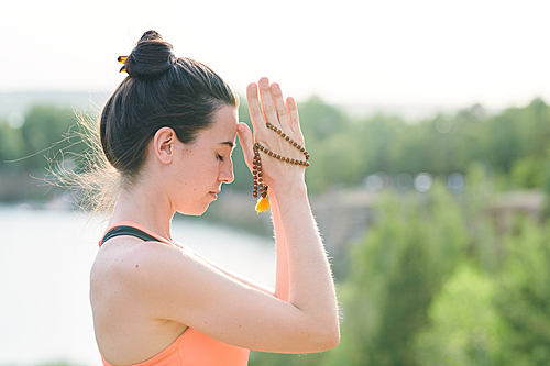 Concentrated young woman with hair bun holding mala beads while performing spiritual practice outdoors