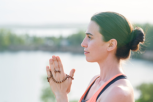 Side view of pensive young woman with hair bun holding mala beads in hands and looking into distance