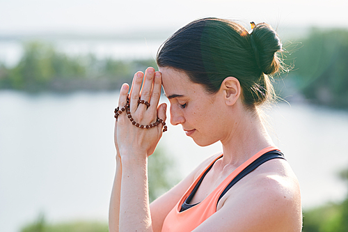 Religious young woman with hair bun joining hands together with beads while praying outdoors