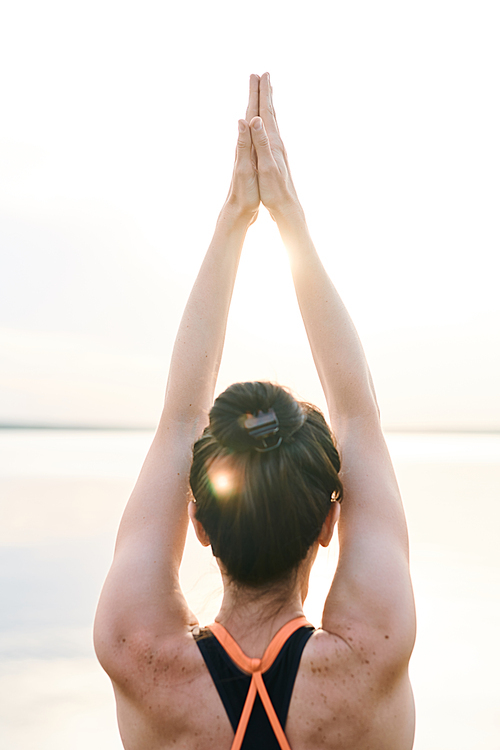 Rear view of girl full of energy standing against sunrise and raising hands up while aligning body with sun salutation exercise