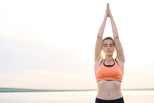 Serious serene young woman in sportswear standing against sea in sunset light and practicing yoga