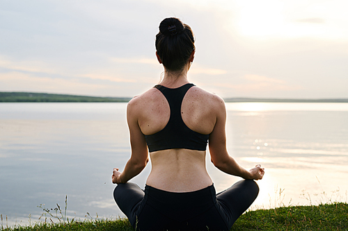 Rear view of young woman in black sportswear sitting with crossed legs on grass and meditating at sunset