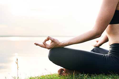 Close-up of unrecognizable woman in leggings sitting on shore of lake and practicing yoga in unity with nature