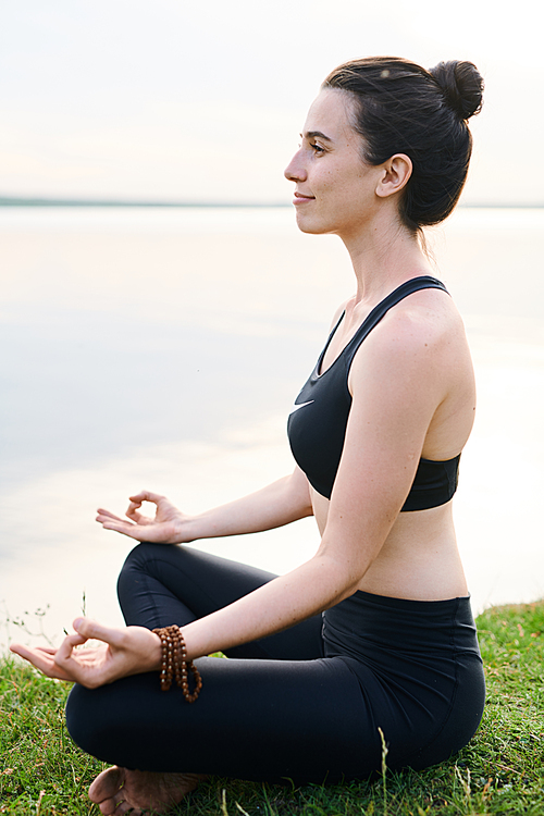 Content attractive woman with hair bun sitting with crossed legs on grass and meditating in open air