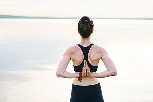 Rear view of young woman with strengthened spine standing against sea and joining hands behind back while practicing yoga in fresh air