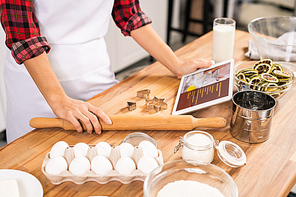 Hands of young housewife holding rolling-pin and tablet with recipe while standing by wooden table and going to cook pastry