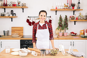Happy young woman in apron standing by table in the kitchen in front of camera while going to make cookies
