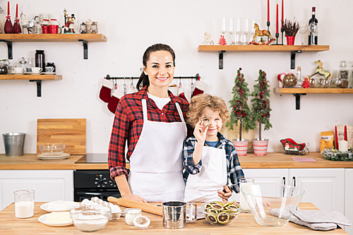 Happy young woman and her little son going to make cookies in the kitchen while standing by table in front of camera