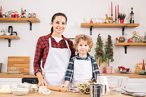 Happy young woman and little boy in aprons preparing pastry in the kitchen while standing by table in front of camera