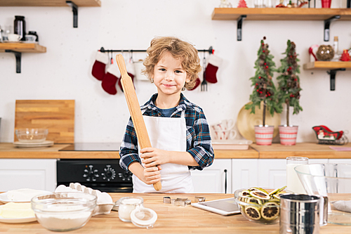Happy little boy in apron holding rolling-pin while going to make cookies in the kitchen to help his mother