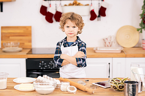 Serious or discontent little boy in apron crossing arms on chest while standing by kitchen table with ingredients for cookies