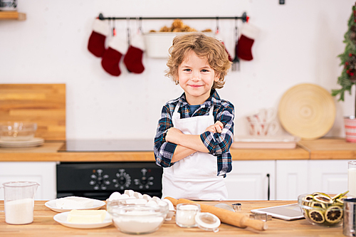 Cheerful curly little boy in apron crossing arms on chest while standing by kitchen table with ingredients for cookies