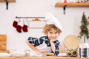 Cute funny little boy in apron and chef hat holding rolling-pin and looking at you while standing by kitchen table with ingredients for cookies