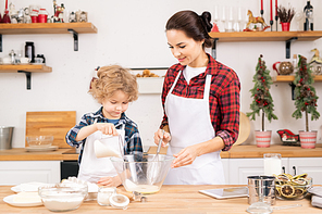 Cute little boy adding sugar into shaked raw eggs in bowl while helping his mother with dough for homemade pastry in the kitchen