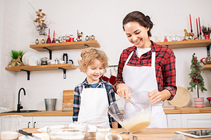 Happy young mother whisking raw eggs in bowl with her little son standing near by while helping his mom