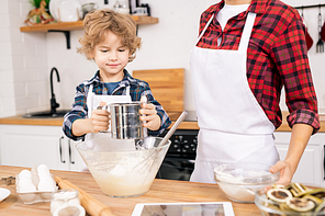 Cute little boy sifting flour while standing by table next to his mom and preparing dough for homemade pastry