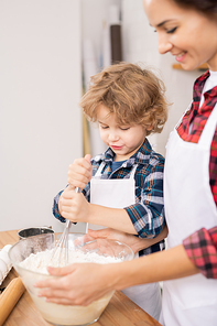 Young female and her son in aprons whisking eggs with flour while preparing dough for homemade cookies at home