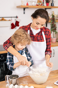 Young female helping her son whisk eggs with flour in bowl while preparing dough for tasty homemade cookies