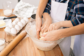 Hands of young woman and her little son kneading selfmade dough in bowl while both standing by table in the kitchen
