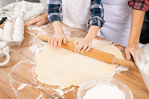 Hands of child with wooden pin rolling fresh dough on table while helping mom with cooking pastry