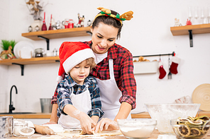 Cute little boy in Santa cap and his mother in Christmas headband making gingerbread cookies in the kitchen