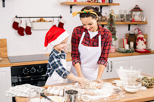 Happy little boy in Santa cap and his mother in Christmas headband kneading dough for cookies in the kitchen