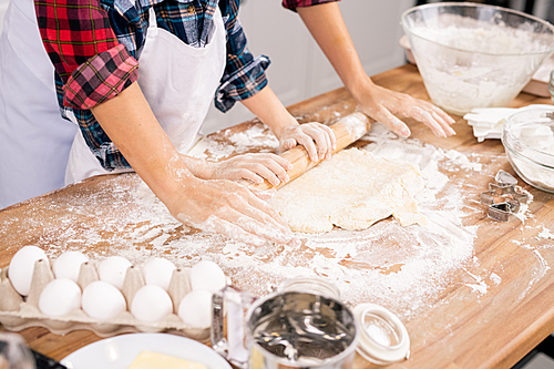 Hands of young female and her son in aprons rolling dough on wooden table while cooking pastry together