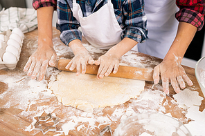 Hands of young female and little boy rolling dough on kitchen table while preparing cookies together