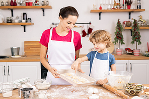 Cute curly boy pointing at one of cookies on tray while discussing its shape with mother in the kitchen