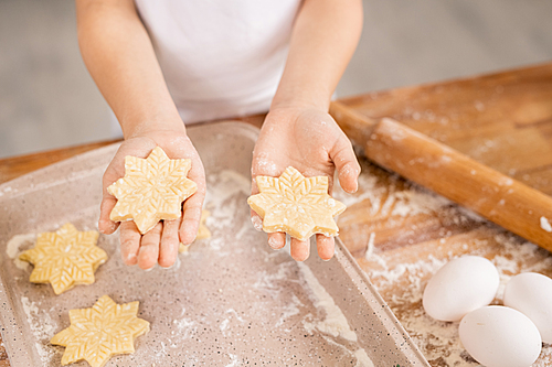 Hands of young woman holding raw snowflake shaped cookies over tray on kitchen table covered with flour