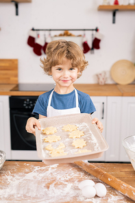 Happy cute boy holding tray with raw selfmade cookies while standing by kitchen table in front of camera
