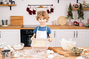 Happy cute boy with raw selfmade cookies looking at you while standing by kitchen table in front of camera