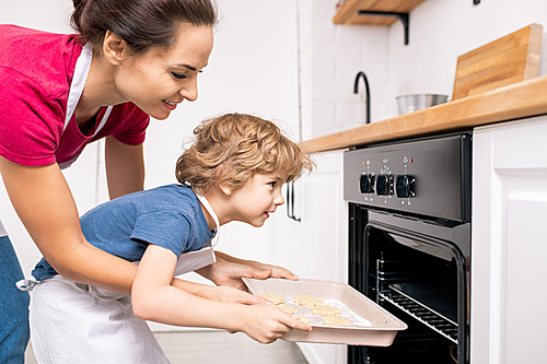 Young female helping her little son put tray with raw cookies into open oven while both bending forwards