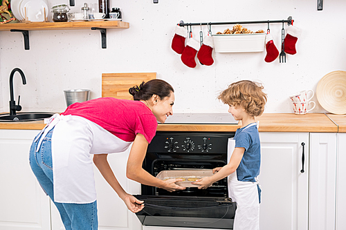 Little boy in apron helping his mom put tray with raw cookies into open oven while both looking at each other