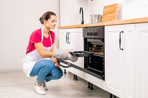 Young housewife in apron squatting while putting tray with raw cookies into electric oven while in the kitchen