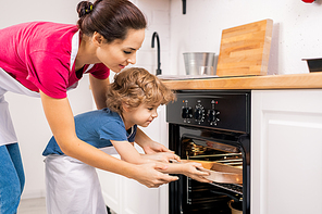 Little boy putting tray into open oven and his mom helping him after making homemade cookies on weekend