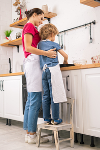 Little boy in apron helping his mom with cooking food while both standing by kitchen table and putting something on tray