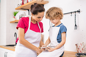 Adorable blond boy in apron holding smartphone while showing his mother new recipe of tasty food in the kitchen